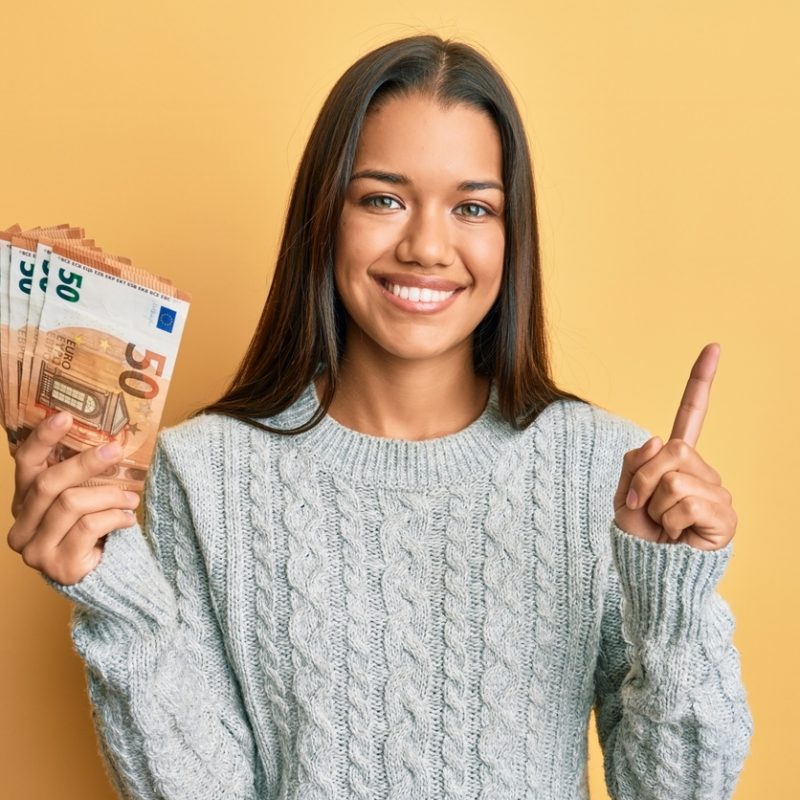 beautiful hispanic woman holding bunch of 50 euro banknotes smiling happy pointing with hand and finger to the side